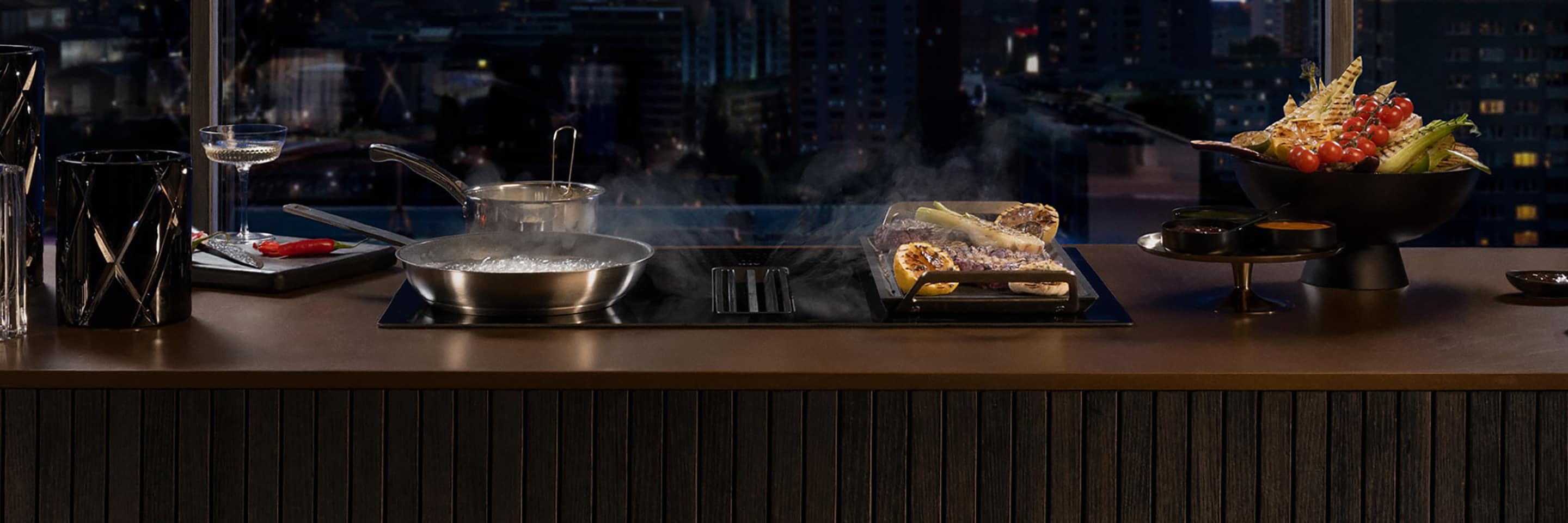AEG Hob2Hood Kochfeld mit integriertem Dunstabzug, Pfanne mit Dampf, City-Skyline-Nacht im Hintergrund